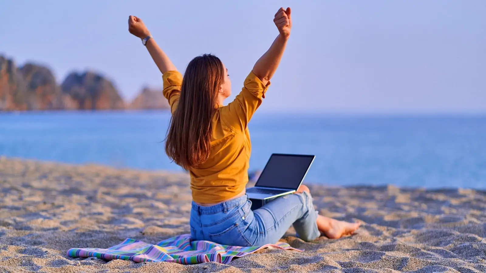 A person working on a laptop while sitting on a beach with a sunset in the background